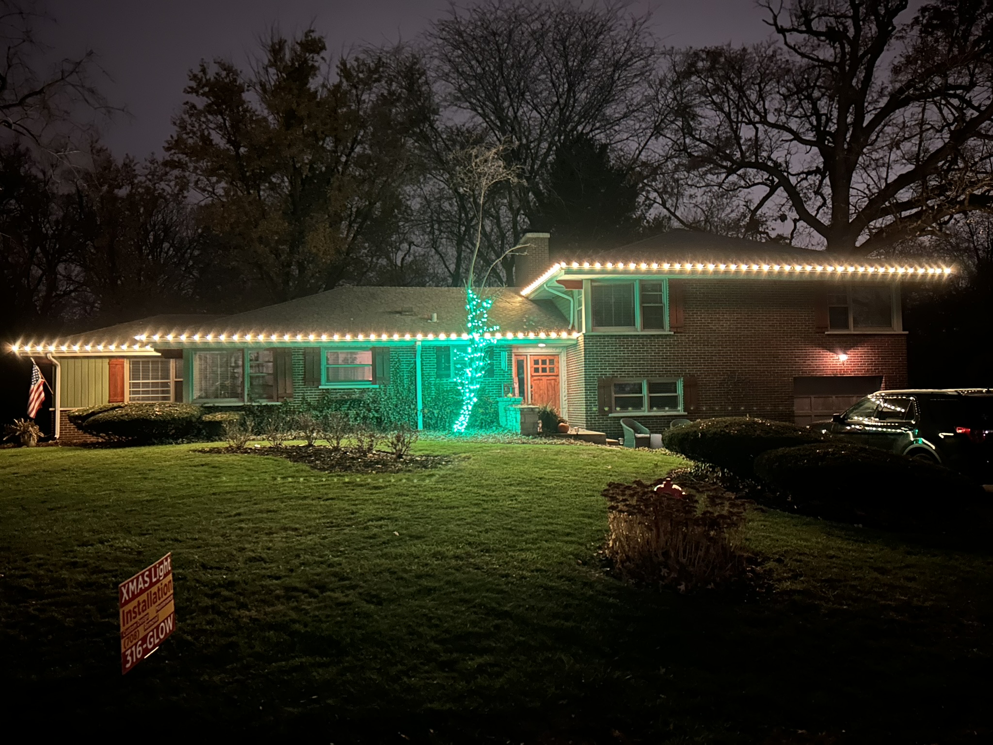 Split-level home with warm white roofline lights and a green wrapped tree